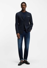 Marynarka w kolorze granatowym o fakturowanej tkaninie, z dwoma guzikami i kieszeniami z patkami, zestawiona z ciemnymi jeansami i czarnymi mokasynami. Poszetka dodaje detali.