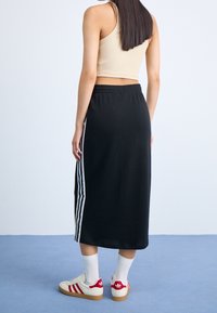 Kvinna som står på blått golv iklädd svart midi kjol med vita ränder, beige crop top, vita strumpor och vita sneakers med röda detaljer.
