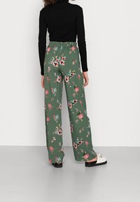 Wijdvallende broek met bloemenpatroon in groen met roze en blauwe bloemen, elastische tailleband, gecombineerd met een zwart shirt met lange mouwen en witte schoenen.