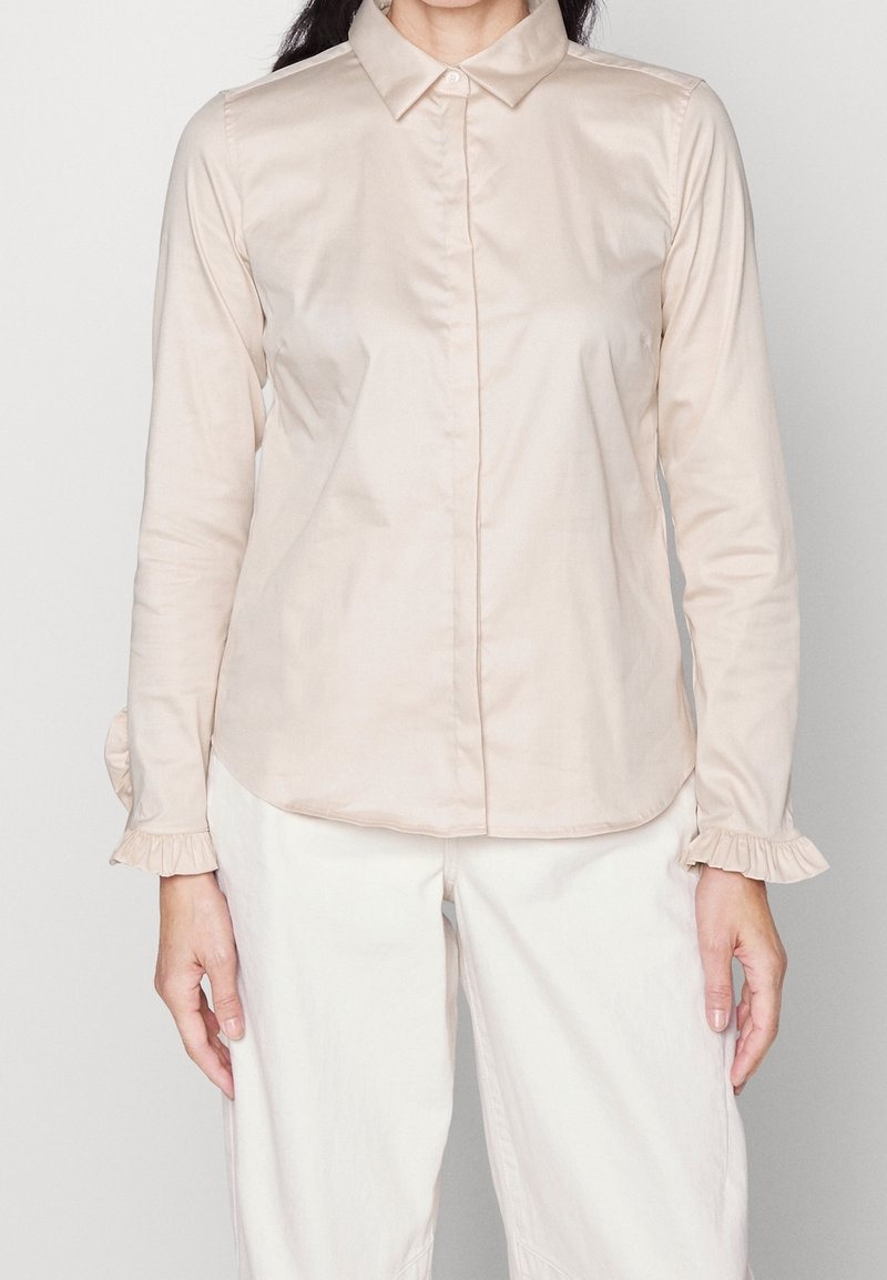 Femme portant un chemisier beige clair à manches longues avec des poignets à volants et un pantalon blanc, debout devant un fond uni.