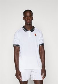 Camisa polo blanca con cuello negro y rayas en las mangas. Presenta un tejido texturizado y un pequeño logo colorido en el lado izquierdo del pecho.