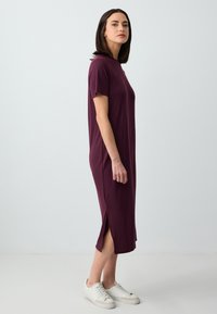 Burgundowa midi sukienka z krótkimi rękawami, bocznymi rozcięciami i miękką teksturą, w połączeniu z białymi trampkami na jednolitym tle.