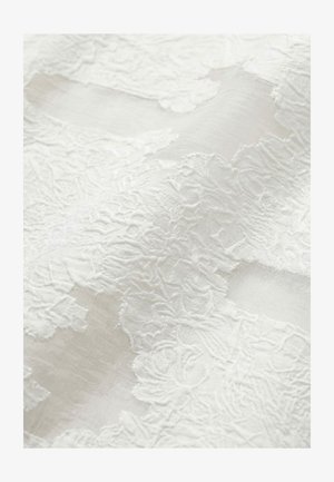 Weißer Stoff mit filigraner floraler Spitzenstickerei, die über durchsichtigem Material liegt und Textur sowie zarte Muster zeigt.