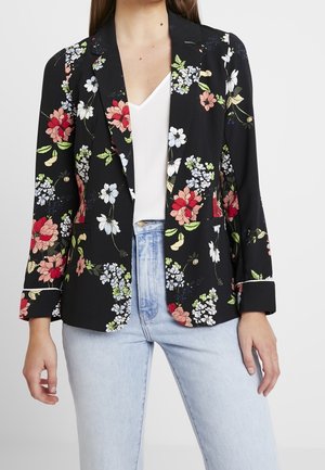 Kvinde iført sort blazer med blomster i rødt, hvidt og grønt, kombineret med hvid top og lyseblå jeans mod en ensfarvet baggrund.