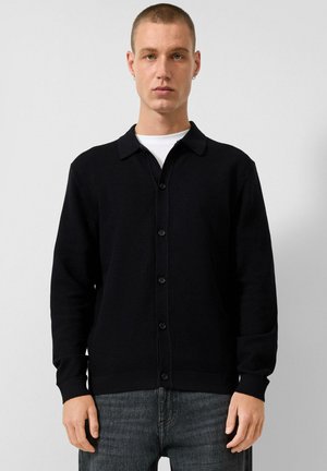 Junger Mann mit kurzen Haaren, der eine schwarze, geknöpfte Strickjacke über einem weißen Hemd und dunklen Jeans trägt, vor einem einfarbigen Hintergrund stehend.