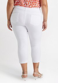 Pantaloni bianchi a vita alta, con tasche posteriori, realizzati in un tessuto liscio. L'orlo è aderente e una maglietta colorata è parzialmente visibile in vita.