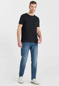 Camiseta negra de manga corta con un bolsillo en el pecho, combinada con jeans azul claro y zapatillas blancas. La tela parece suave con un corte estándar.