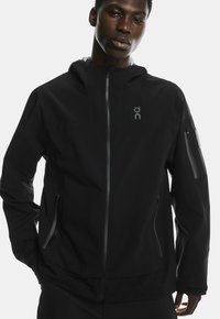 Černá voděodolná bunda s kapucí, která má kapsy na zip a reflexní akcenty. Hladký povrch s moderním designem.