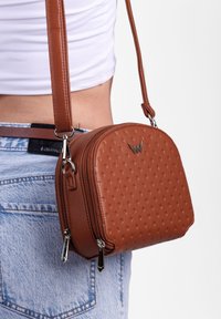 Hnědá kožená crossbody taška s texturovanými tečkami a stříbrnými detaily. Kulatý tvar, dvě uzavíratelné přihrádky a nastavitelné ramenní pouzdro.