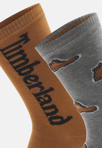 Due paia di calze: una arancione con il logo "Timberland" in nero e una grigia con grafiche di stivali marroni. Entrambe hanno polsini a coste e una texture liscia.