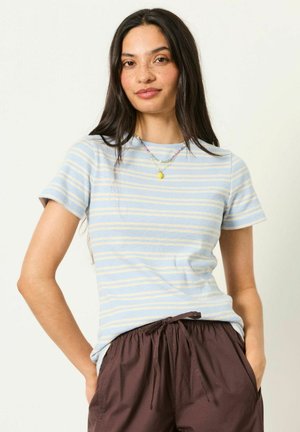 Jonge vrouw met lang donker haar die een lichtblauw en geel gestreept T-shirt met korte mouwen en donkerbruine broek met koord draagt.