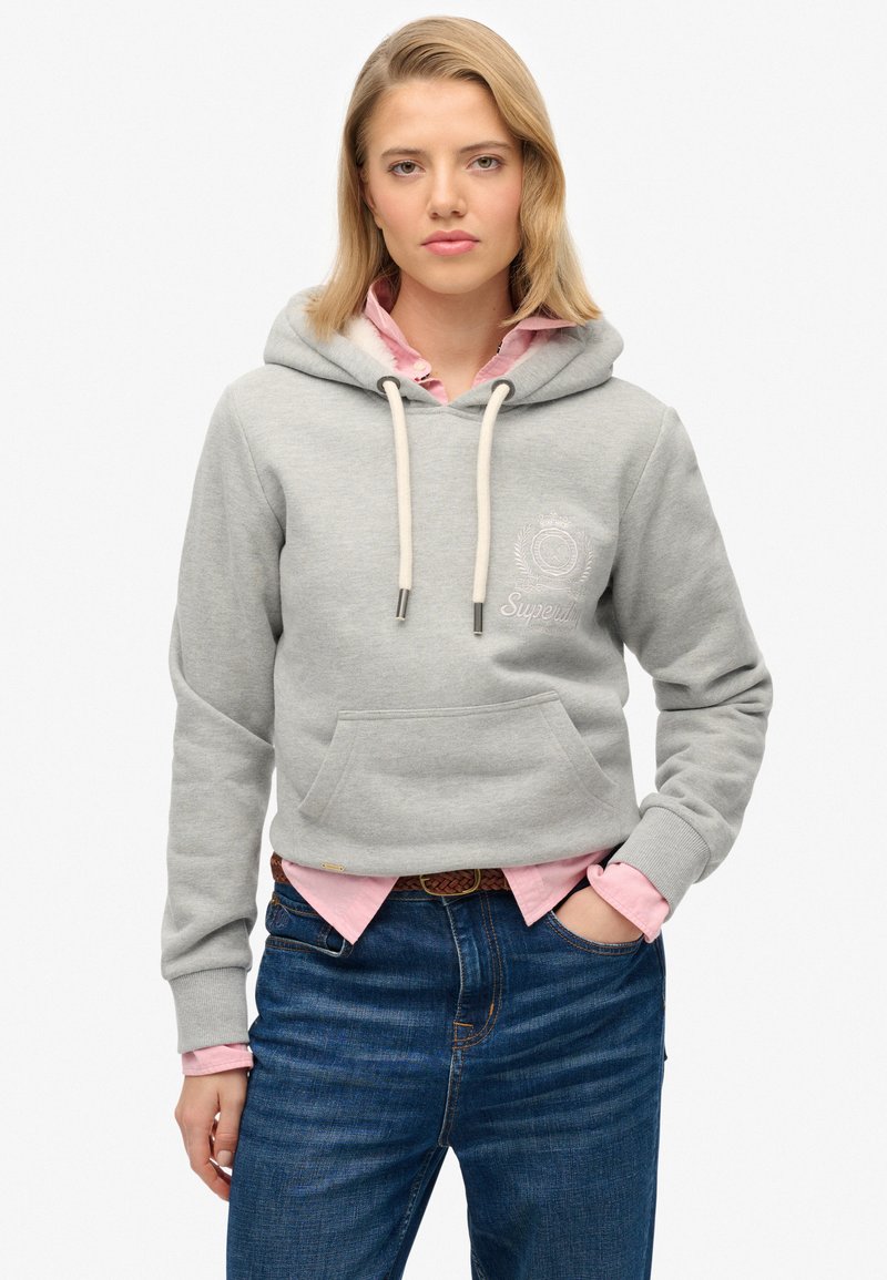 Sudadera gris con bolsillo frontal, capucha con cordón y logo bordado. Lleva puesta una camisa de color rosa con cuello y está combinada con jeans azules.