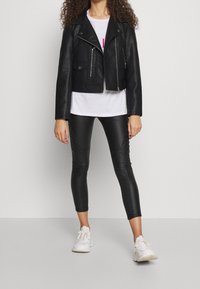 Kvinna iklädd en svart läderjacka, vit t-shirt, svarta vadderade läderleggings och vita sneakers som står mot en neutral bakgrund.