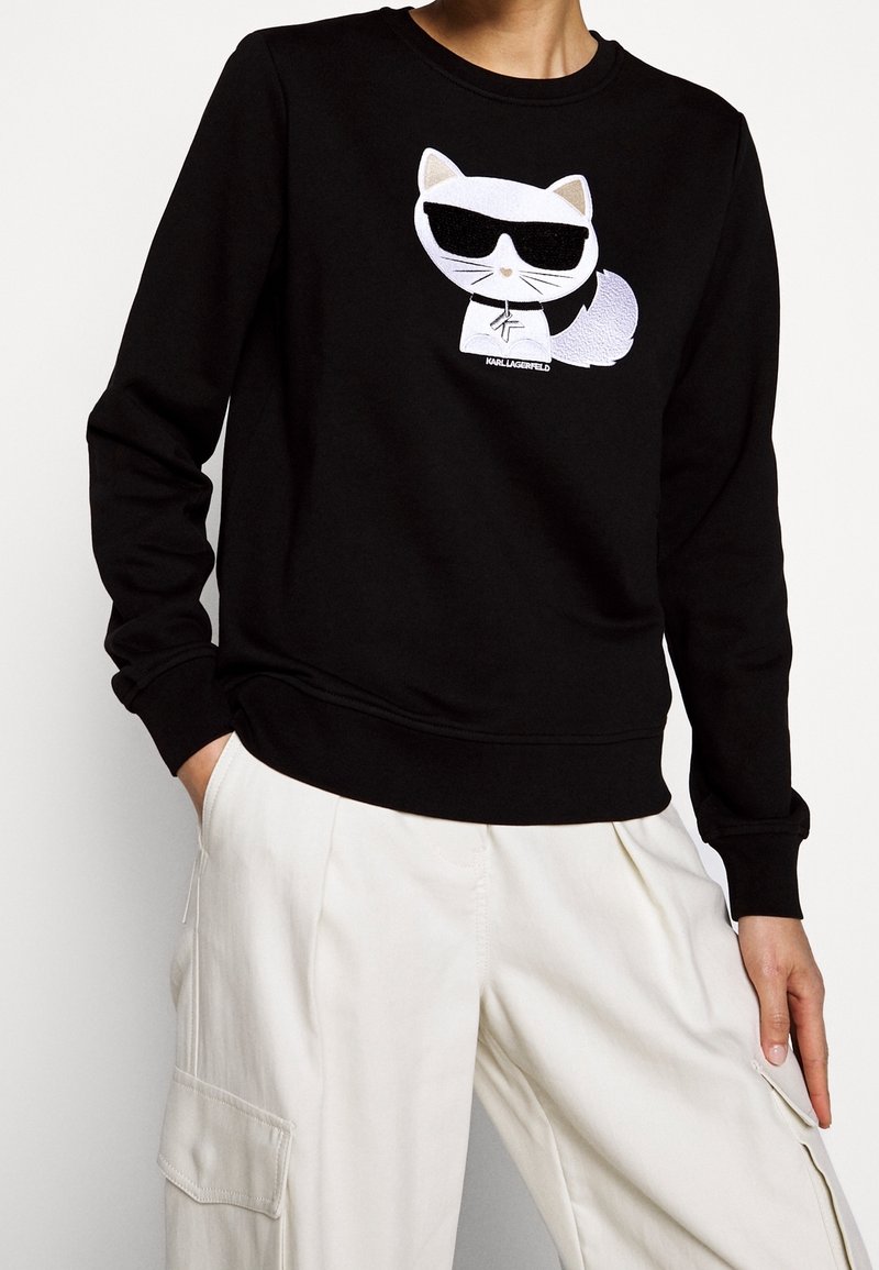 Sudadera negra con un gráfico de un gato de dibujos animados en blanco que usa gafas de sol. El diseño incluye detalles bordados y un cuello redondo.