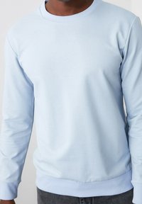Sweat-shirt bleu clair à col rond en matériau doux et lisse. Dispose de manches longues et de poignets et d'un ourlet côtelés pour un look ajusté.