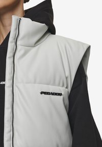 Lys grå ermeløs puffer vest med høy krage, glidelås foran, med en svart logo, lagd over en svart hettegenser.