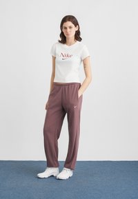 Jovem mulher de pé com as mãos nos bolsos, vestindo uma t-shirt branca da Nike, calças de fato de treino cor malva e ténis brancos em um tapete azul.