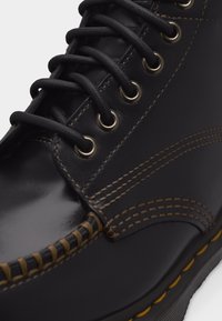 Μαύρο δερμάτινο παπούτσι με καφέ ραφές, μαύρα στρογγυλά κορδόνια περασμένα μέσα από μεταλλικά κουμπώματα και υφασμένο άκρο σόλας με κίτρινες λεπτομέρειες.