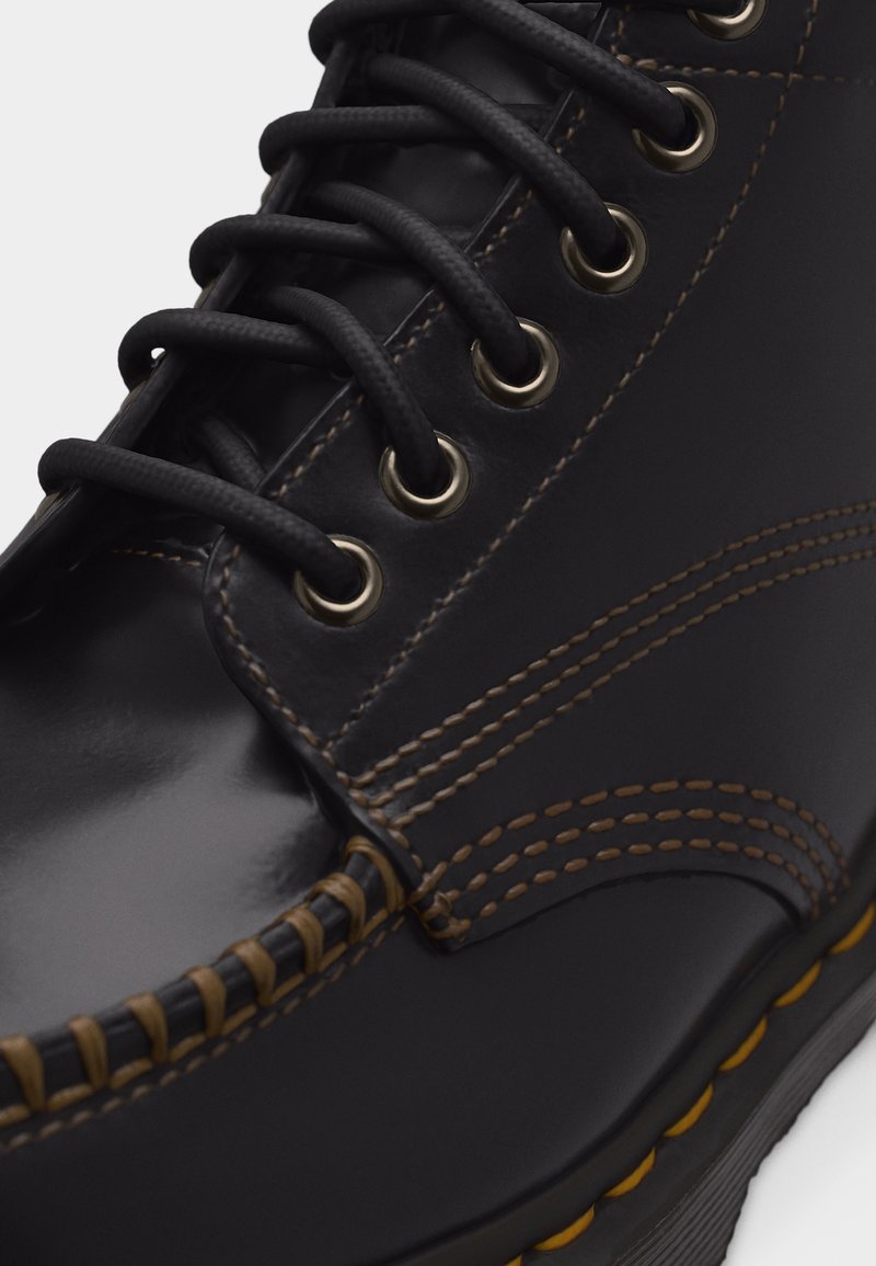 Zwarte leren schoen met bruine stiksels, zwarte ronde veters die door metalen oogjes zijn geregen, en een getextureerde zoolrand met gele accenten.