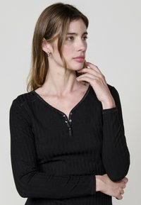 Haut noir à manches longues côtelé avec un décolleté en V et patte de boutonnage à trois boutons. Texture douce avec un subtil éclat et des détails de surpiqûres contrastées.