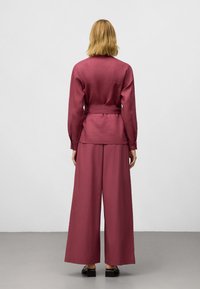 Mörkröd tvådelad outfit som består av en långärmad topp med knytband i midjan och vida byxor, tillverkad av ett slätt tyg med minimal textur.