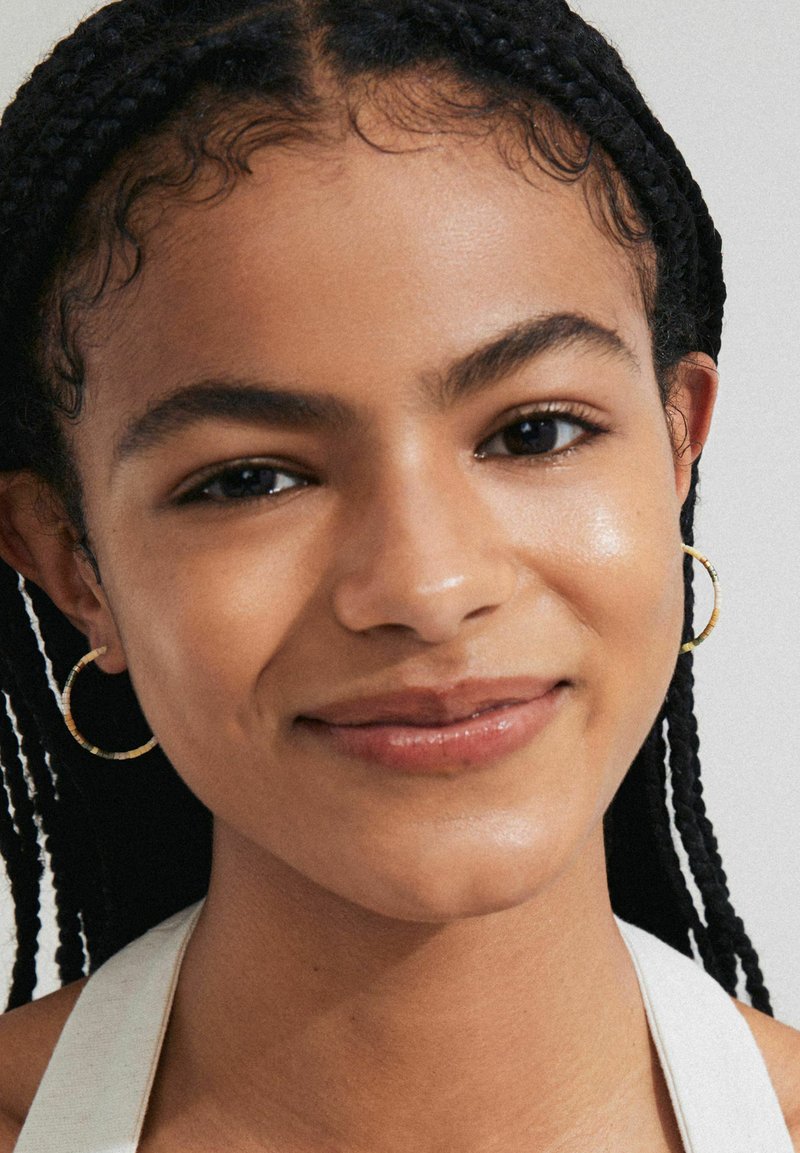 Close-up of a person with braided hair, wearing small gold hoop earrings, and a white top. Smooth skin with a subtle glow, neutral expression.