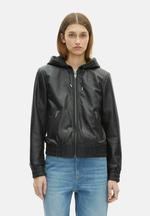 Femme portant une veste en cuir noire à capuche avec fermeture éclair et poches zippées, associée à un jean bleu clair, sur fond blanc.