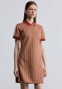 Rozsdaszínű pólóruha geometriai mintával, rövid ujjakkal és gallérral. Logó díszítéssel és kontrasztos oldalsávokkal.