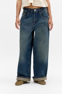 Person med vide blå jeans med rullekanter, beige mokasiner og en cropped off-white topp, som står mot en hvit bakgrunn.