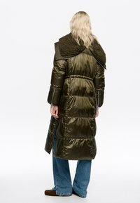 Puszysty oliwkowozielony długi płaszcz z fakturowanym ciemnozielonym kołnierzem, wcięciem w talii i poziomym pikowaniem; noszony na niebieskie dżinsy i brązowe buty.