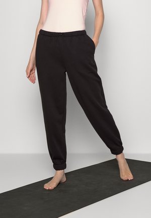 Black sweatpants with an elastic waistband, side pockets, and tapered cuffs, made of soft fabric, worn with a light pink top on a yoga mat.