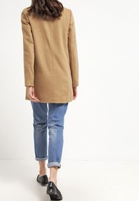 Béžový vlněný kabát se střihem do rovna, dlouhými rukávy a minimalistickým designem, kombinovaný s světle modrými stočenými džíny a černými nazouvacími botami.