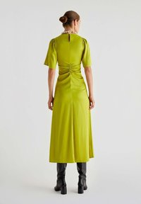 Abito midi in satin verde con maniche a sbuffo corte, dettaglio arricciato sul retro, scollatura a occhiello e una silhouette fluida, abbinato a stivali neri con tacco.