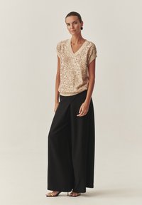 Bēša spīguļojoša augšdaļa ar V veida kakla izgriezumu un īsām piedurknēm, kombinēta ar melnām platām biksēm. Gluda tekstūra ar brīvu piegriezumu.