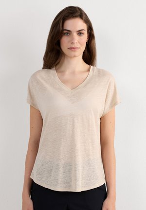 Femme aux longs cheveux bruns portant un haut beige clair à manches courtes et col en V, debout devant un fond blanc uni.