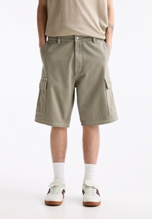 Homme portant un short cargo beige, des chaussettes blanches montantes et des baskets blanches avec des bandes sombres, debout les mains dans les poches sur fond blanc.