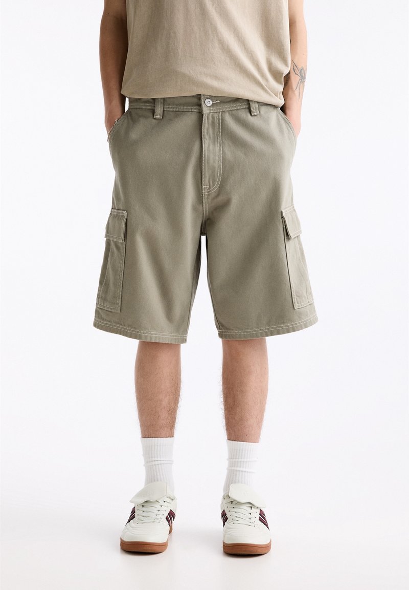 Homme portant un short cargo beige, des chaussettes blanches montantes et des baskets blanches avec des bandes sombres, debout les mains dans les poches sur fond blanc.