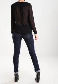 Blouse noire transparente à manches longues avec un col rond, associée à un jean denim foncé ajusté et des bottines. Textures minimalistes et lisses.