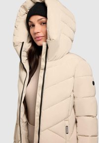 Vrouw in een beige gewatteerde jas met grote capuchon en zwarte beanie, kijkend naar de camera tegen een effen achtergrond.