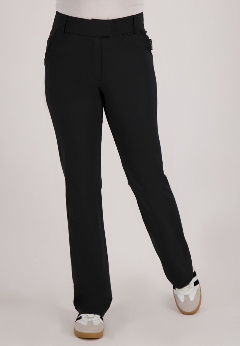 Persoon draagt zwarte rechte broek en witte sneakers, staat met één been gekruist voor het andere tegen een effen achtergrond.