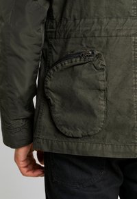 Olivgrön jacka med en zip-funktionen cirkel ficka, texturerad tyg och synliga sömnadsdetaljer på axlarna och sidorna.