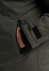 Olijfgroene jas met een ritszak, zwarte geribbelde manchetten en een zachte, gestructureerde stof. Inclusief klittebandaccenten bij de mouw.