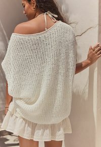 Bijela pletena oversized veste s otvorenim tkanjem, uparena s krem suknjom s višim slojevima koja ima vezene akcentne detalje i rupičaste elemente.
