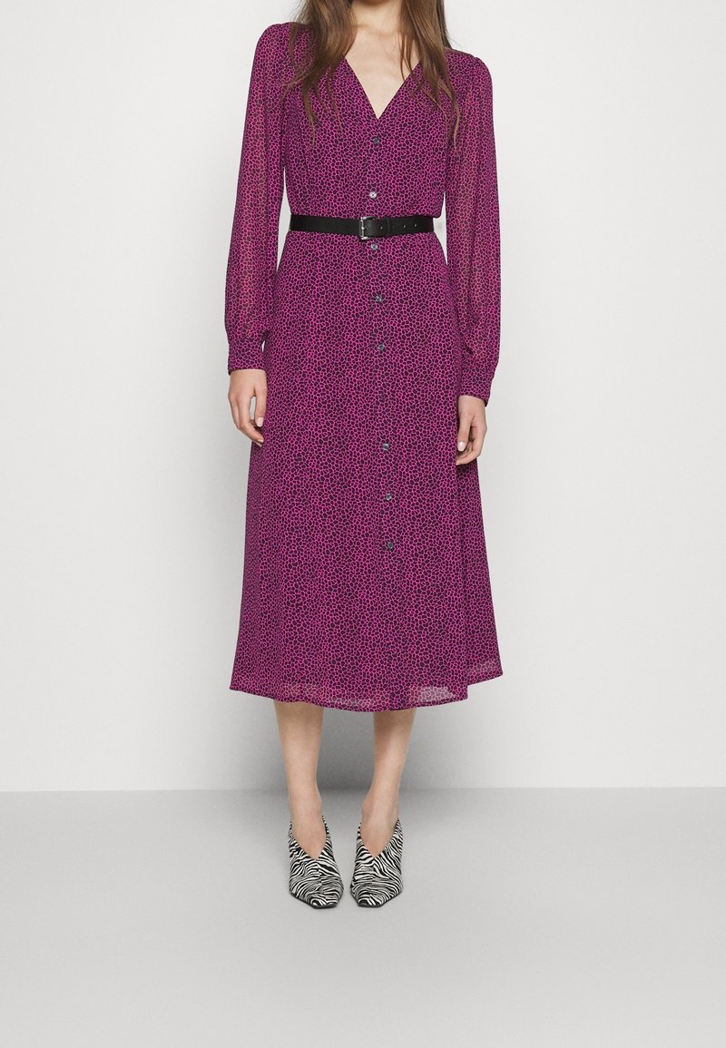 Femme portant une robe midi violette à manches longues avec des taches noires, une ceinture noire et des chaussures pointues à rayures zébrées, debout devant un fond uni.