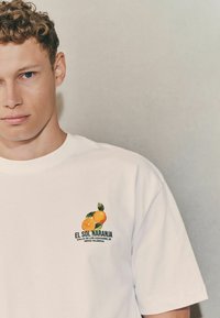 White cotton t-shirt featuring a colorful orange fruit graphic with green leaves and text "EL SOL NARANJA" on the left chest area.