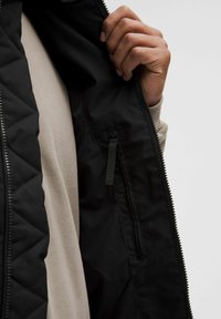 Schwarze gesteppte Jacke, die geöffnet ist und ein beiges Innenfutter zeigt. Verfügt über eine seitliche Tasche mit Reißverschluss und eine glatte Stoffoberfläche.