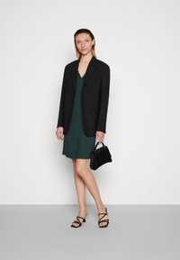 Blazer preto sobre um vestido verde com decote em ondas; modelo segura uma bolsa preta e usa sandálias de salto pretas com tiras. Fundo simples.