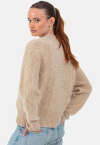 Pull beige en maille torsadée avec motifs texturés, ourlet côtelé et coupe ample. Tissu doux et col montant pour plus de chaleur et de confort.