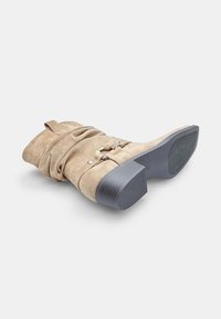 Beige velúr bokacsizma laza dizájnnal, ezüst színű fém részletekkel és vastag fekete sarkakkal. Texturált talp a tapadásért.