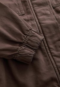 Manche de veste marron avec poignet élastique et couture visible sur tissu épais, vue rapprochée du haut du bras et de la zone de l'épaule.